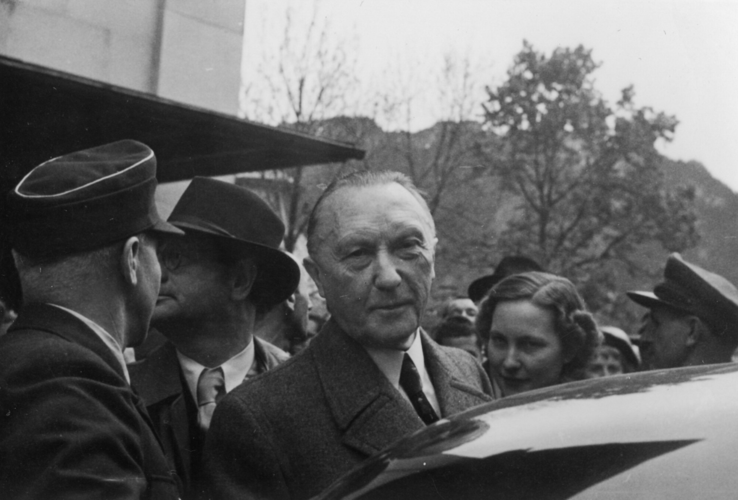 Adenauer vor einem Auto in die Kamera schauend