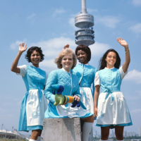 Hostessen in blau-weißen Kleidern mit Maskottchen Waldi vor dem Olympiaturm.
