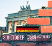 Symboldbild. Bild einer kaputten Mauer mit deutscher Flagge vor dem Brandenburger Tor.