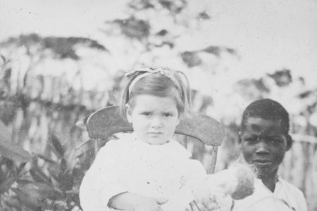 Kind aus Kolonialmacht in weiß gekleidet, erhöht vor einer Einheimischen sitzend.